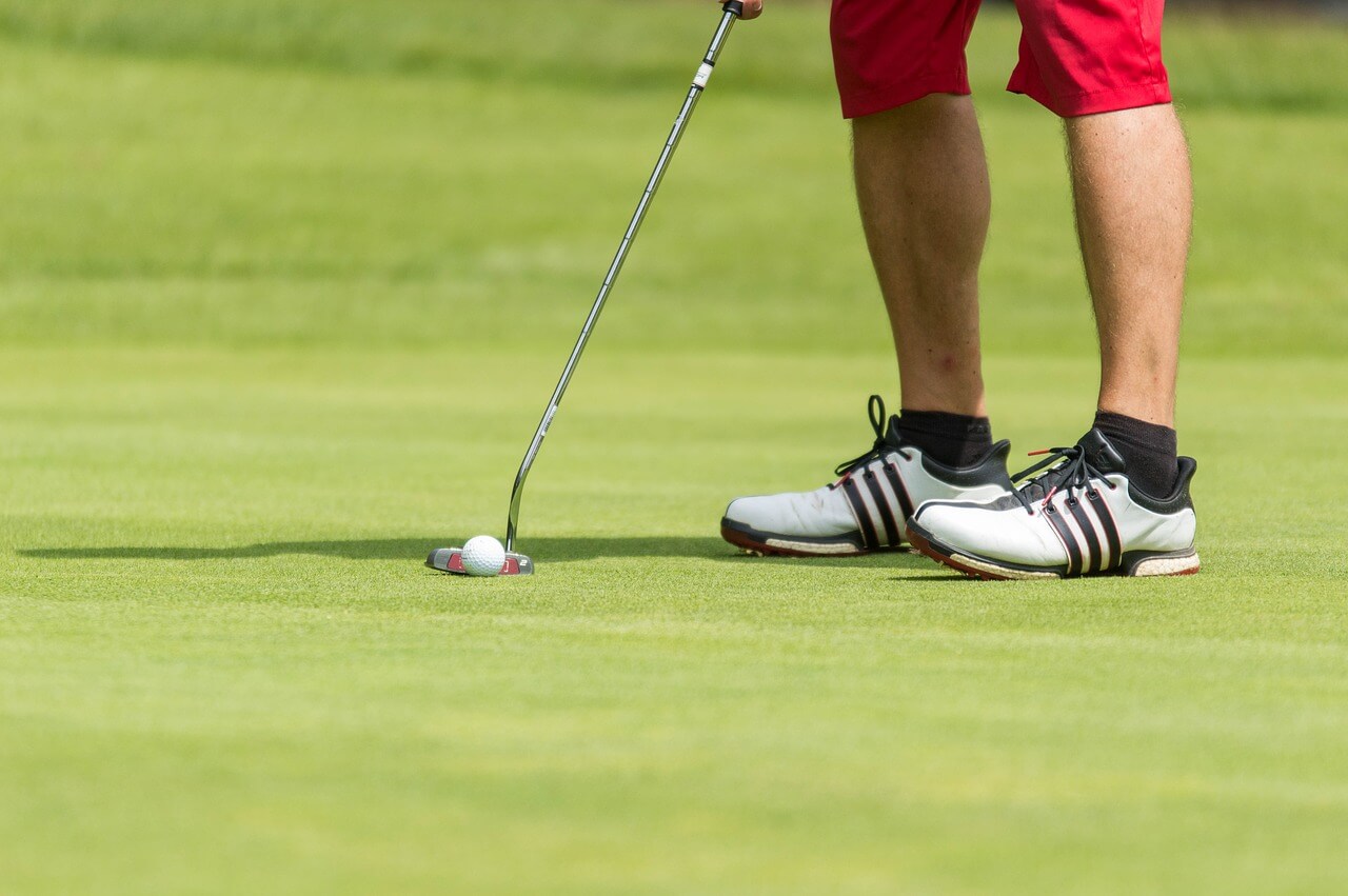 Man hitting a golf ball wearing white shoe with black stripes and red shorts