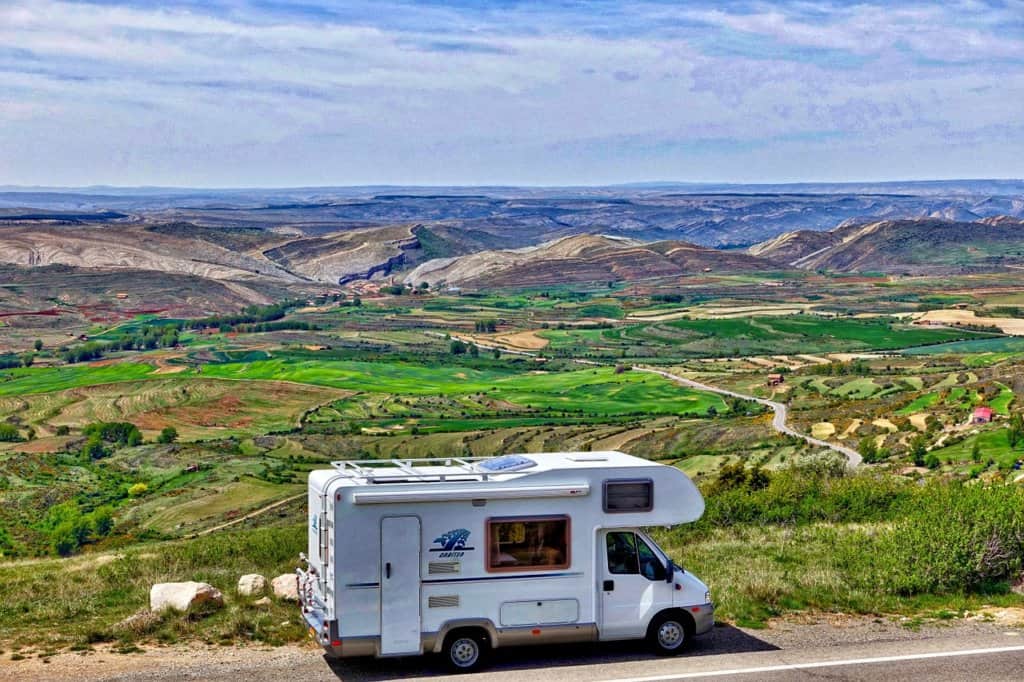 A white RV driving past a large green gorge with a blue sky 