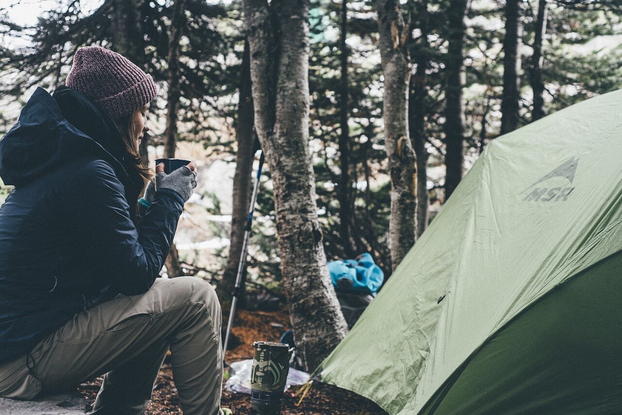 Woman sitting in the woods next to a tent holding a cup of coffee