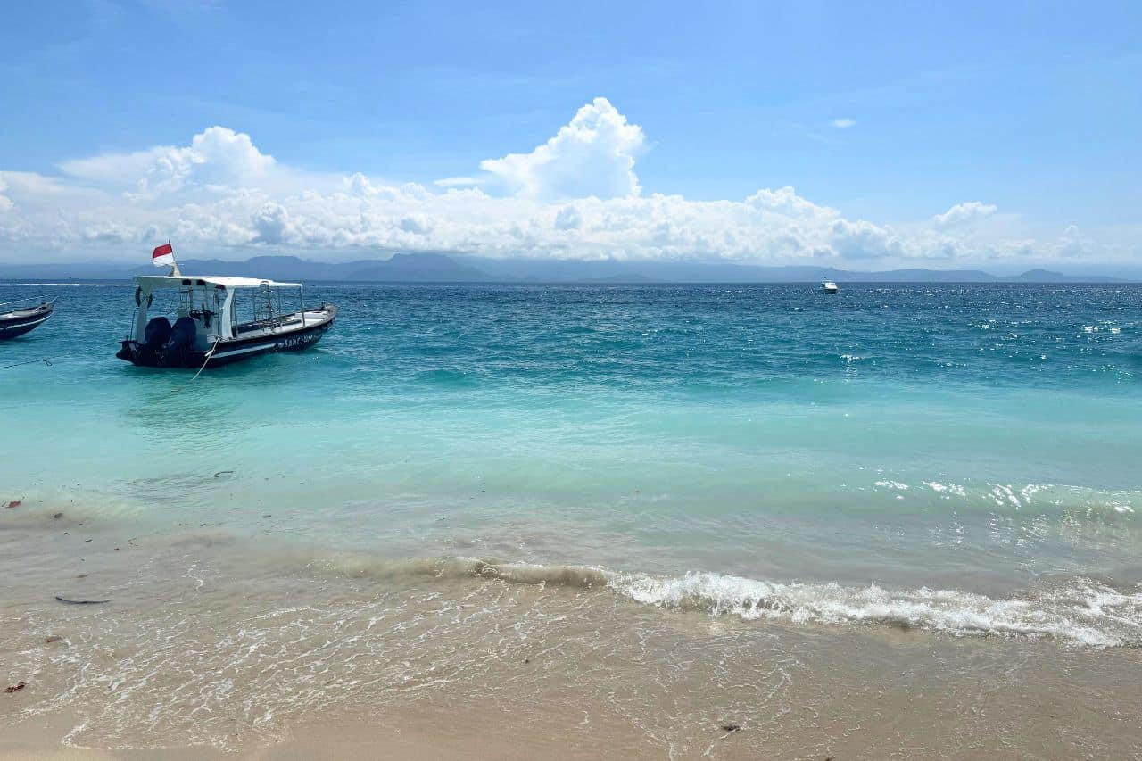 A small boat floating on turquoise waters near a sandy beach in Bali with mountains visible in the distance.
