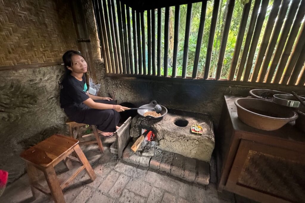 A Balinese woman roasting coffee beans over a wood fire in a traditional bamboo hut.