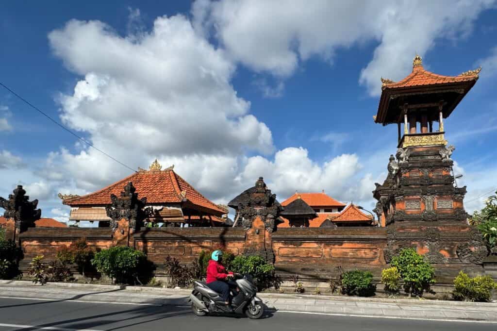 Scooter rider passing a traditional Balinese temple with orange tiled roofs on a sunny day.