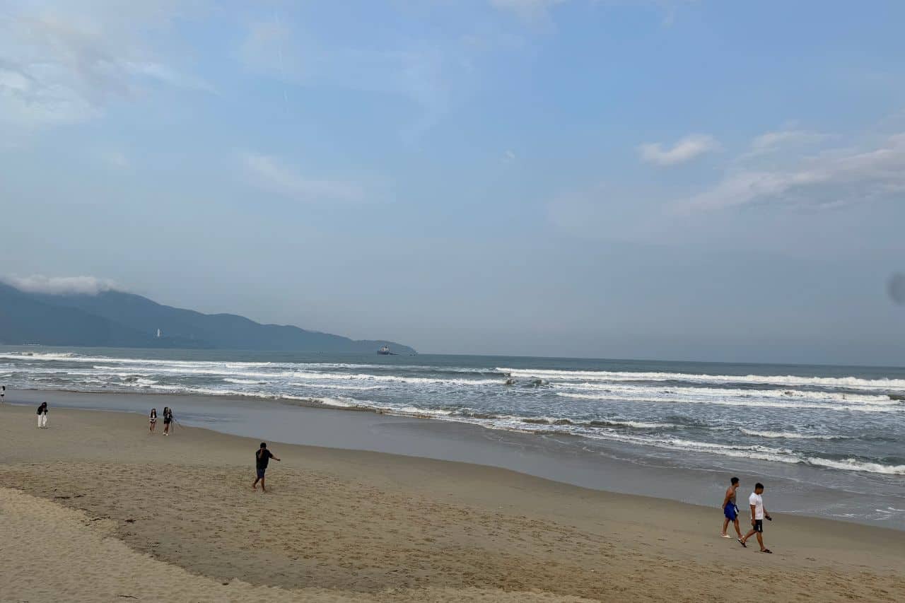 People walking along the shoreline at My Khe Da Nang Beach with mountains in the background