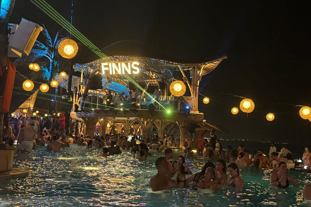 Nighttime pool party at Finns Beach Club with people swimming and dancing under hanging lights.