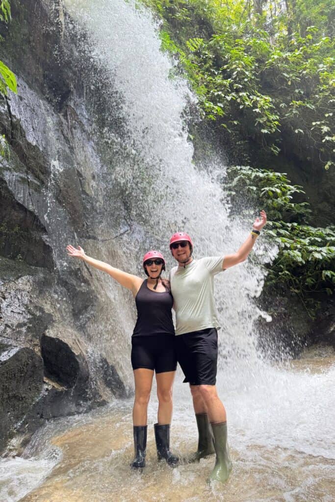 Smiling couple wearing helmets and boots standing in front of a jungle waterfall during an Ubud adventure tour