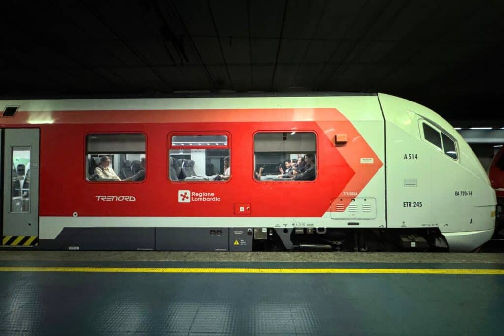 Side view of a red and white Trenord train with passengers seated, at a station platform