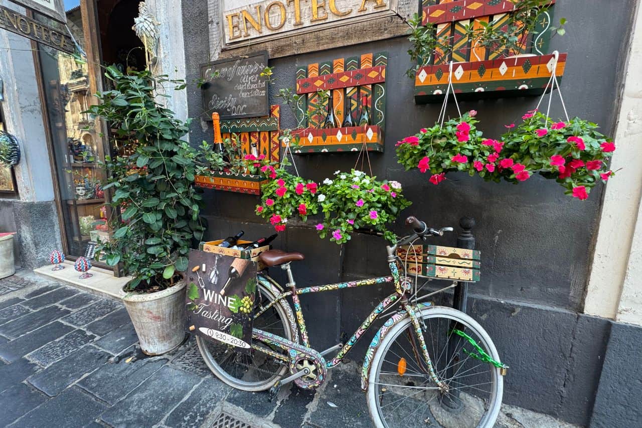 A colorful bike sits in front of a Catania wine store, surrounded by vibrant flowers, capturing Sicilian charm.
