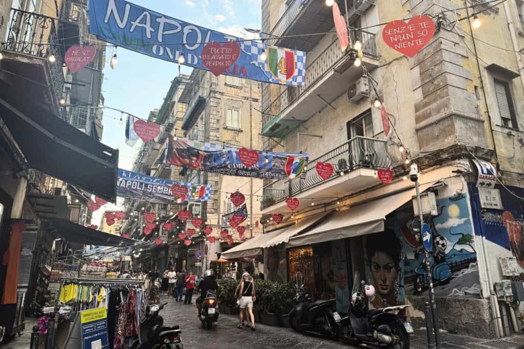 Narrow Naples street decorated with blue and white “Napoli Sempre” banners hanging overhead that you will see during your 1 day in Naples Italy