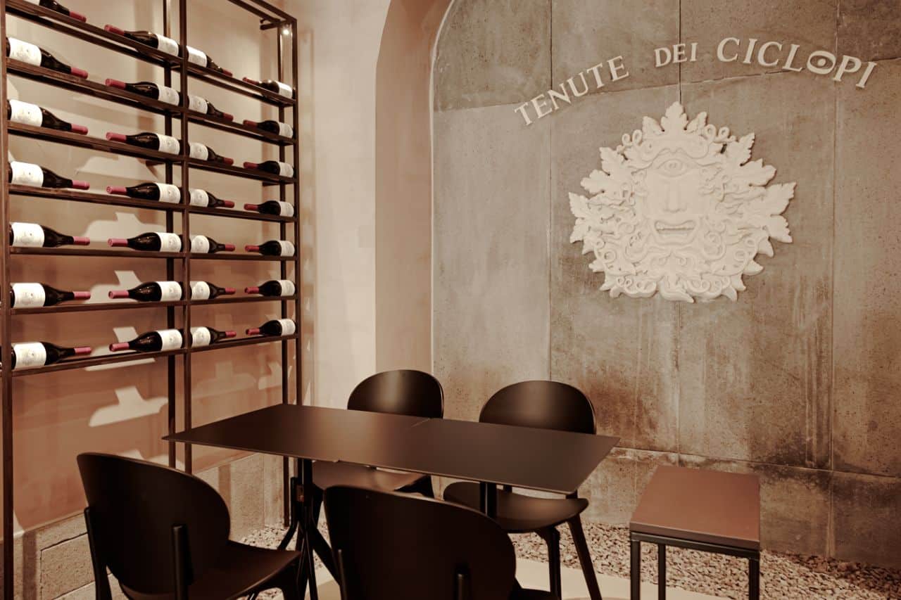 Seating area inside the Tenute dei Ciclopi wine tasting room in Catania with Etna wine bottles displayed on the wall.